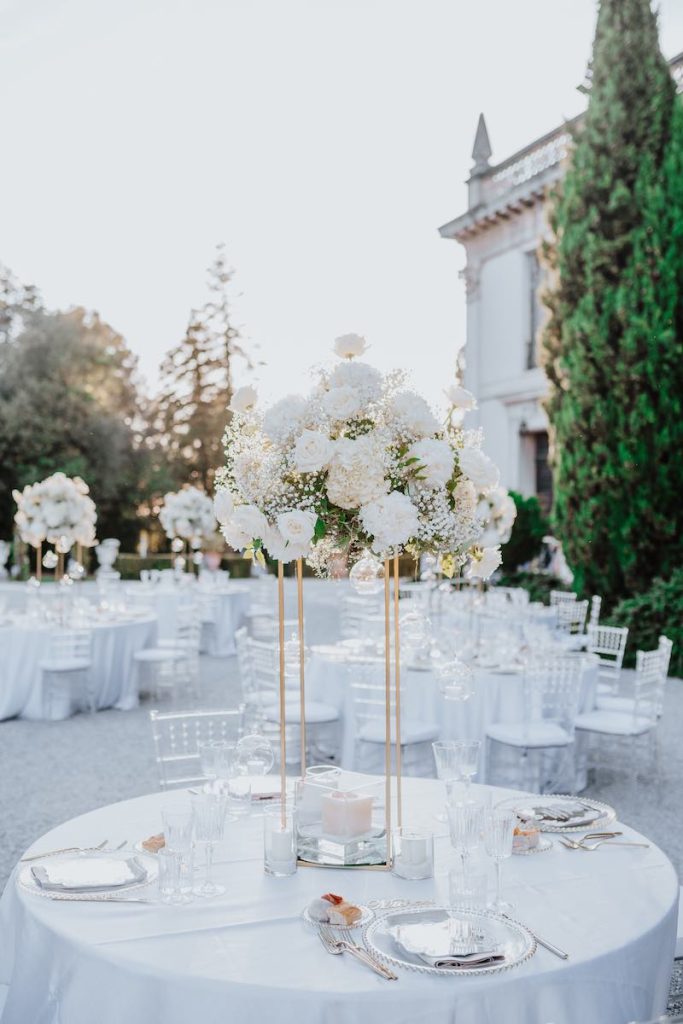 In questa foto un tavolo matrimonio rotondo allestito nello stile d Tatiana con un alzata dorata con fiori di colore bianco, piatti di vetro trasparente e stoviglie argento