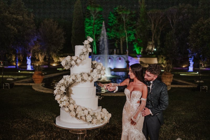 In questa foto due sposi di un matrimonio di Tatiana Tabanelli mentre tagliano una torta a piani decorata con un elica di rosa di colore bianco