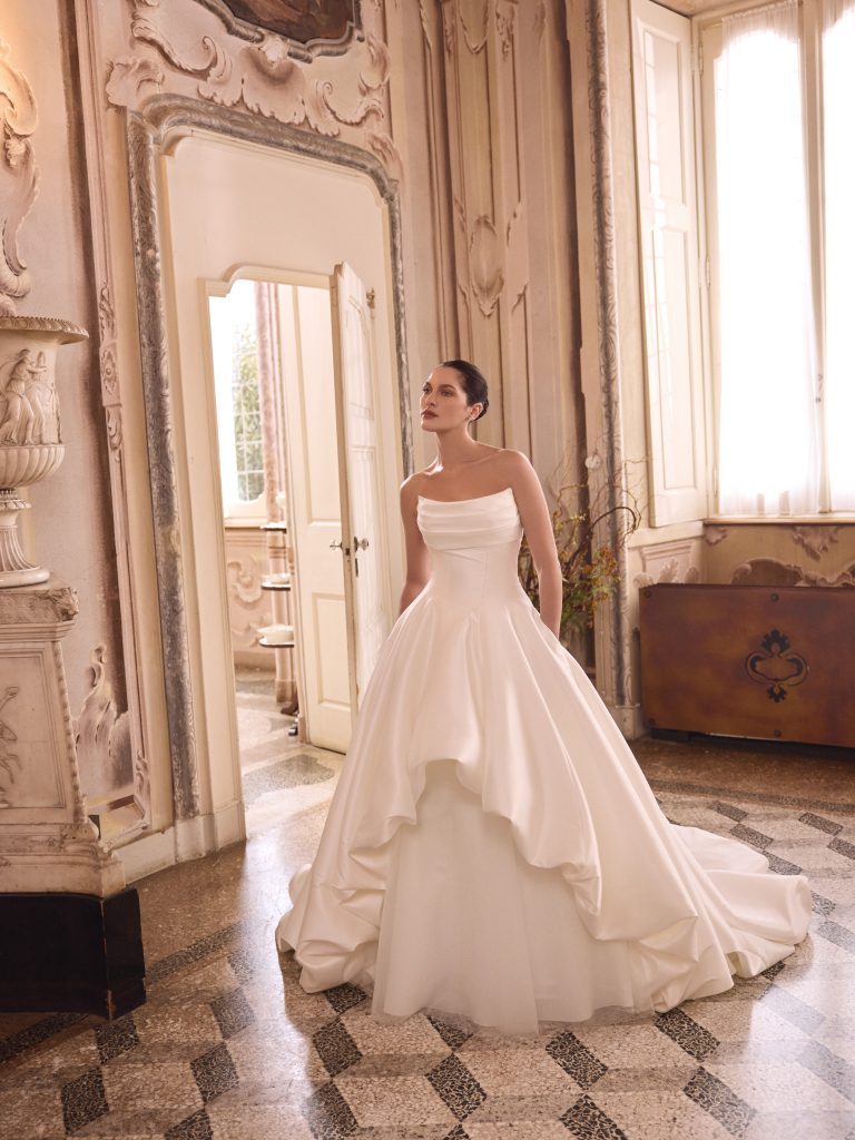 In questa foto, una sposa indossa un abito elegante in mikado dalla linea principesca, con corpino drappeggiato e ampia gonna asimmetrica. Lo scatto, ambientato in una sala d’epoca con pareti decorate e pavimento in marmo, valorizza il fascino intramontabile degli abiti da sposa particolari 2026.