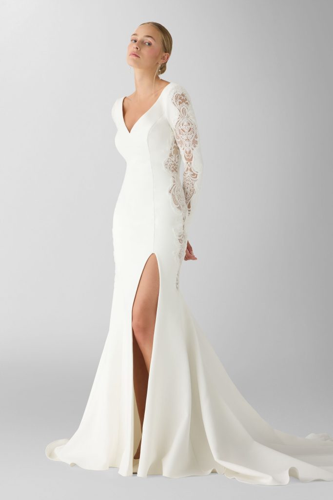In questa foto, una modella indossa un abito bianco a sirena con profondo spacco frontale e maniche lunghe in pizzo floreale. Il design è pulito ma ricco di dettagli raffinati, perfetto per valorizzare una figura slanciata. Un esempio sofisticato di abiti da sposa invernali 2026, che unisce eleganza e sensualità con gusto minimal.