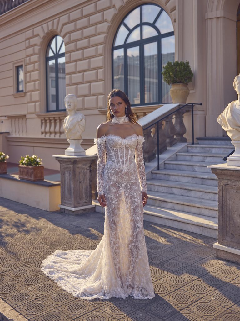 In questa foto, una modella indossa un abito da sposa particolare 2026 firmato Michela Ferriero, posando davanti a una villa aristocratica. Il vestito, interamente in pizzo trasparente con dettagli preziosi, ha maniche lunghe, un corpetto a bustier e un alto collo di pizzo. Il design romantico e audace trasmette un’eleganza teatrale.