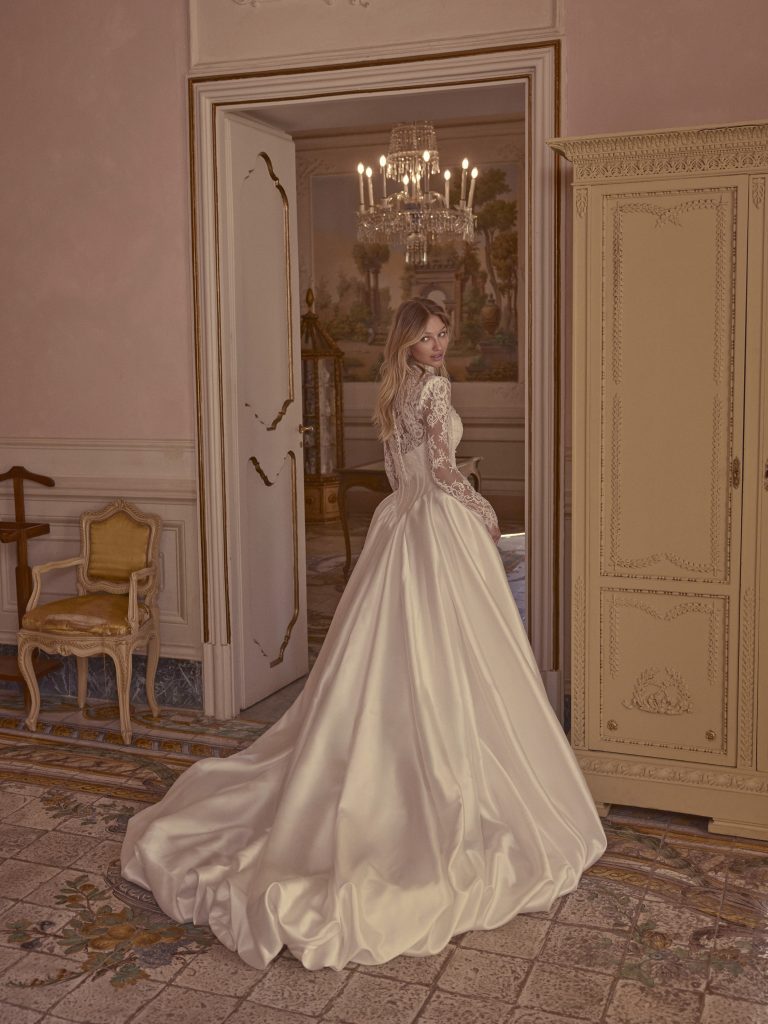In questa foto, una sposa si volta mostrando l’ampio strascico di un abito in raso lucido color avorio. Il corpetto in pizzo trasparente con maniche lunghe e la scena d'interni barocchi creano un’atmosfera sontuosa. Un’opzione romantica e regale tra gli abiti da sposa invernali 2026.