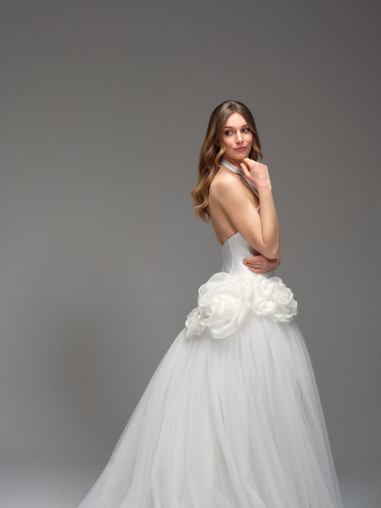 In questa foto, una modella guarda verso la fotocamera con un sorriso discreto, indossando un abito da sposa moderno 2026 con corpino liscio e gonna in tulle vaporoso. Ai fianchi, l’abito presenta due grandi rose in tessuto, che aggiungono un tocco contemporaneo e scultoreo al look.