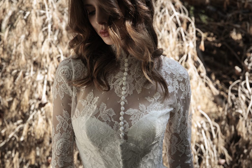 In questa foto ravvicinata, il dettaglio del corpetto in pizzo floreale con bottoncini ricoperti enfatizza la delicatezza del design. I capelli sciolti e la luce naturale rendono l’immagine poetica. È un’elegante rappresentazione della raffinatezza negli abiti da sposa invernali 2026.