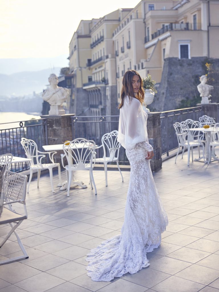 In questa foto, una sposa posa su una terrazza con vista mare, indossando un abito in pizzo a sirena con strascico e una mantella leggera e fluida. La luce calda e l’atmosfera romantica esaltano il fascino degli abiti da sposa invernali 2026.