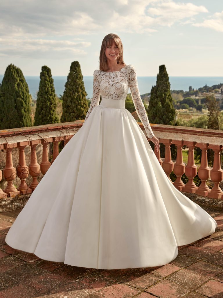 In questa foto, una sposa sorride su una terrazza affacciata sul mare, indossando un abito principesco con gonna voluminosa in mikado e corpetto in pizzo trasparente a maniche lunghe. Lo sfondo mediterraneo esalta la luminosità dell’abito, perfetto per i matrimoni da sogno tra gli abiti da sposa invernali 2026.