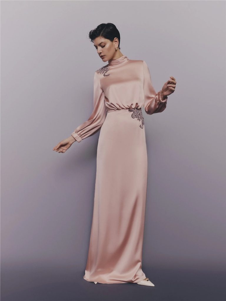 In questa foto una modella posa indossando uno degli abiti da cerimonia Pronovias 2026 di colore rosa cipria, modello scivolato a collo alto con ricami 3D gioiello sulla spalla e sul punto vita