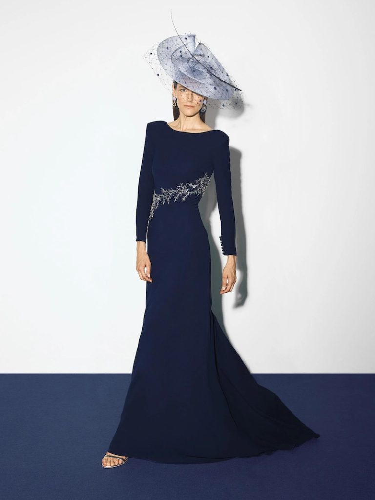 In questa foto una modella posa indossando un abito da cerimonia Pronovias 2026 di colore blu notte, modello a semi sirena con maniche lunghe, scollo a barchetta e dettaglio gioiello sul punto vita. La modella indossa anche un cappello a falda larga di colore turchese con veletta