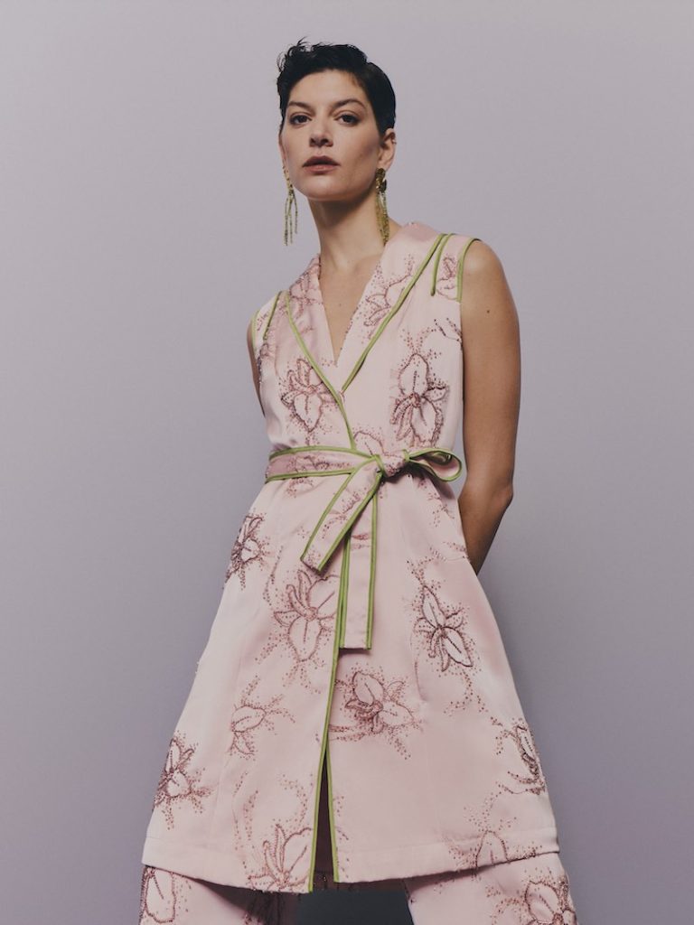 In questa foto una modella posa indossando uno degli abiti da cerimonia Pronovias 2026 di colore rosa cipria con ricami di fiori tono su tono, composto da gilet lungo con revers e cintura abbinata e pantaloni