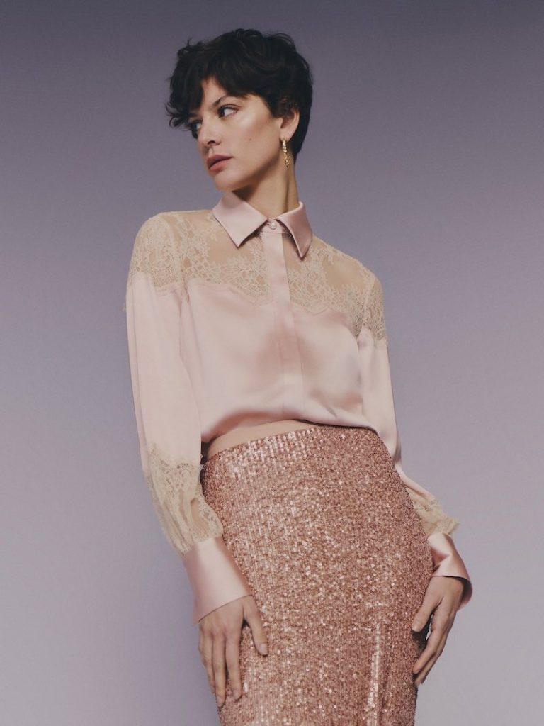 In questa foto una modella posa indossando uno degli abiti da cerimonia Pronovias 2026 di colore rosa cipria composto da camicia in seta e pizzo e una gonna longuette con paillettes