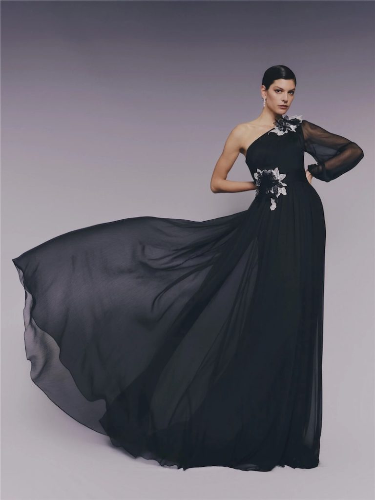 In questa foto una modella posa indossando un abito da cerimonia Pronovias 2026 di colore nero, modello scivolato, monospalla, con fiori 3D, lungo strascico e un manica balloon