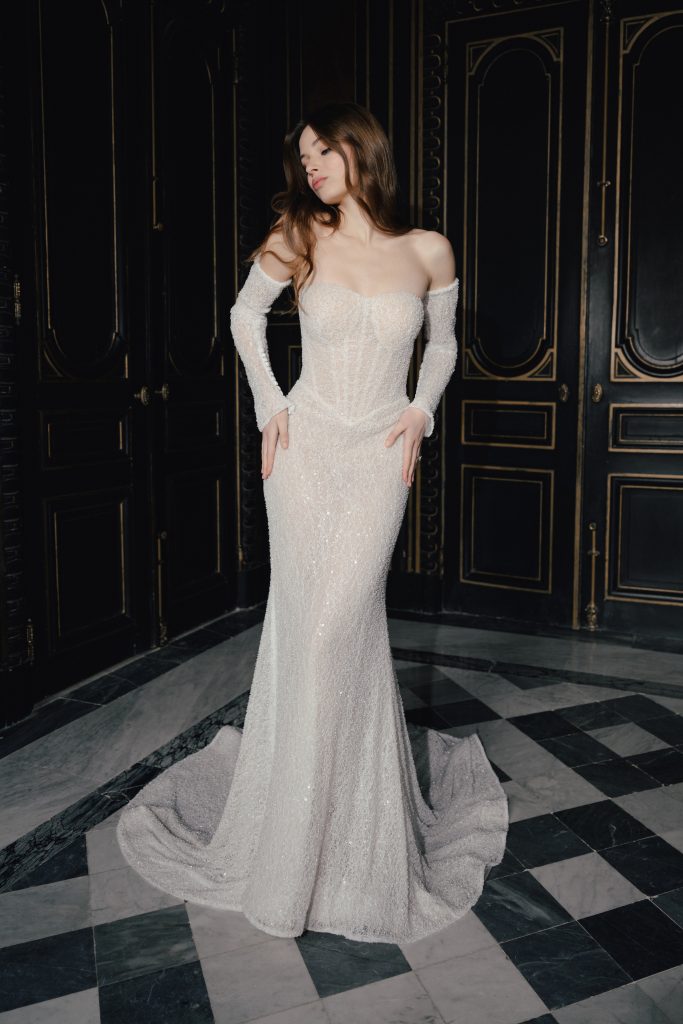 In questa foto, una modella indossa un elegante abito da sposa con spalle scoperte e maniche lunghe trasparenti, interamente ricoperto di paillettes luminose, in una sala dalle pareti scure e ornate. L'immagine fa parte della collezione di abiti da sposa invernali 2026 di Allure Bridals