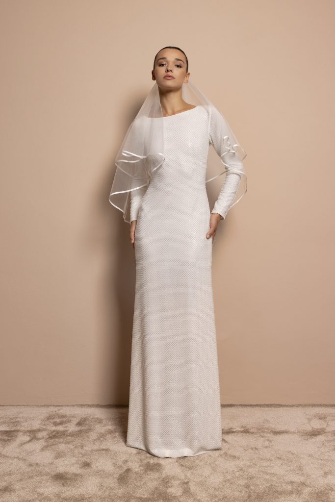 In questa foto, una sposa posa su fondo neutro indossando un abito dritto, interamente ricamato con micro perline tono su tono. Il modello ha maniche lunghe, scollo asimmetrico e un velo corto con profili ondulati. Una scelta raffinata e contemporanea tra gli abiti da sposa invernali 2026.