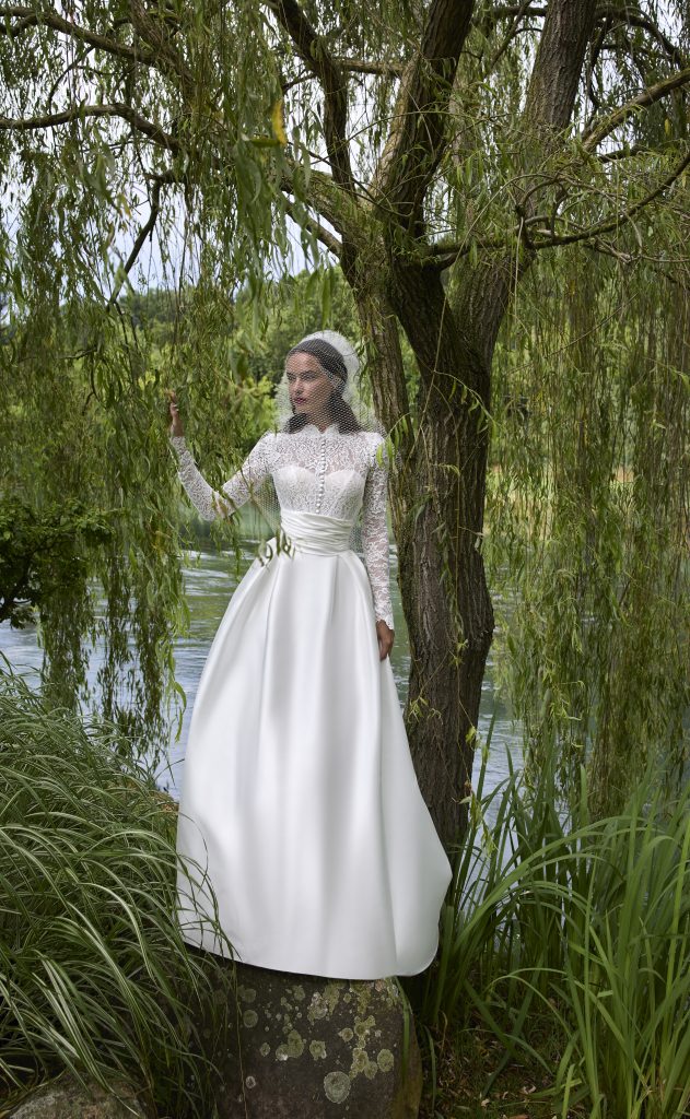 In questa foto, una modella posa all’aperto accanto a un albero, indossando un elegante abito da sposa invernale 2026. La parte superiore in pizzo è arricchita da maniche lunghe e un corpetto velato, mentre la gonna in mikado scivola ampia e lucente. Il velo corto in rete aggiunge un tocco romantico e retrò alla scena immersa nella natura.