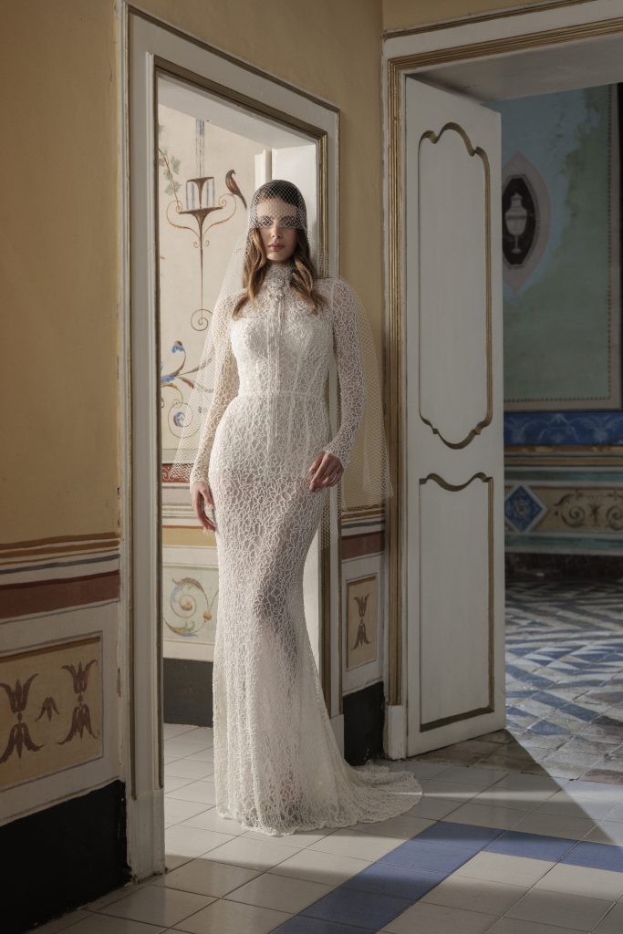 In questa foto, una modella indossa un abito da sposa invernale 2026 dallo stile vintage e raffinato. L’abito è interamente in pizzo con maniche lunghe e collo alto, aderente e trasparente nei punti giusti. La modella si trova in una dimora storica, in piedi sotto una porta antica, e completa il look con un velo corto a rete che le copre il viso.