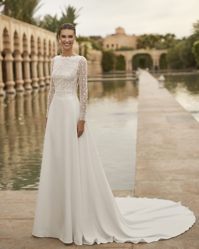 In questa foto, una sposa sorride indossando un abito da sposa bianco con maniche lunghe in pizzo e una gonna semplice e fluida, mentre si trova accanto a una lunga vasca d’acqua in un ambiente esterno ispirato all’architettura araba. L’immagine rappresenta uno degli abiti da sposa invernali 2026 di Rosa Clarà