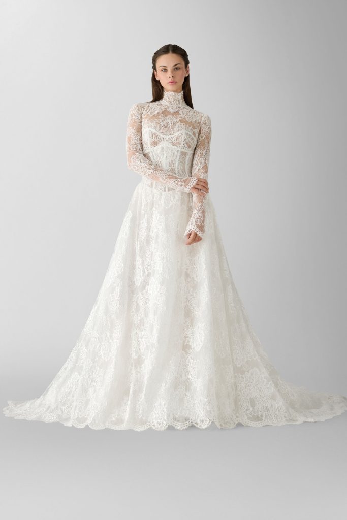 In questa foto, una modella è ritratta in posa frontale con le mani incrociate. Indossa un abito in pizzo a collo alto con maniche lunghe trasparenti e gonna ampia, in perfetto stile romantico. Il design raffinato esprime al meglio l’estetica degli abiti da sposa invernali 2026.