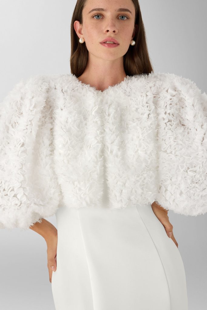 In questa foto, una modella indossa un capospalla voluminoso in tessuto bianco effetto piumato, con maniche ampie a tre quarti e texture tridimensionale. L’outfit, elegante e minimale, fa parte delle tendenze degli abiti da sposa invernali 2026.