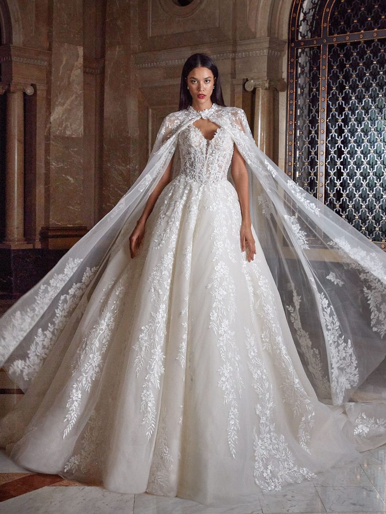 In questa foto, una sposa sfila con un sontuoso abito da sposa invernale 2026 in un ambiente regale. Il vestito è ampio, decorato con motivi floreali in pizzo e abbinato a un lungo mantello trasparente che ricade sulle braccia e sulle spalle. L'effetto è teatrale e maestoso, perfetto per chi sogna un matrimonio da favola.