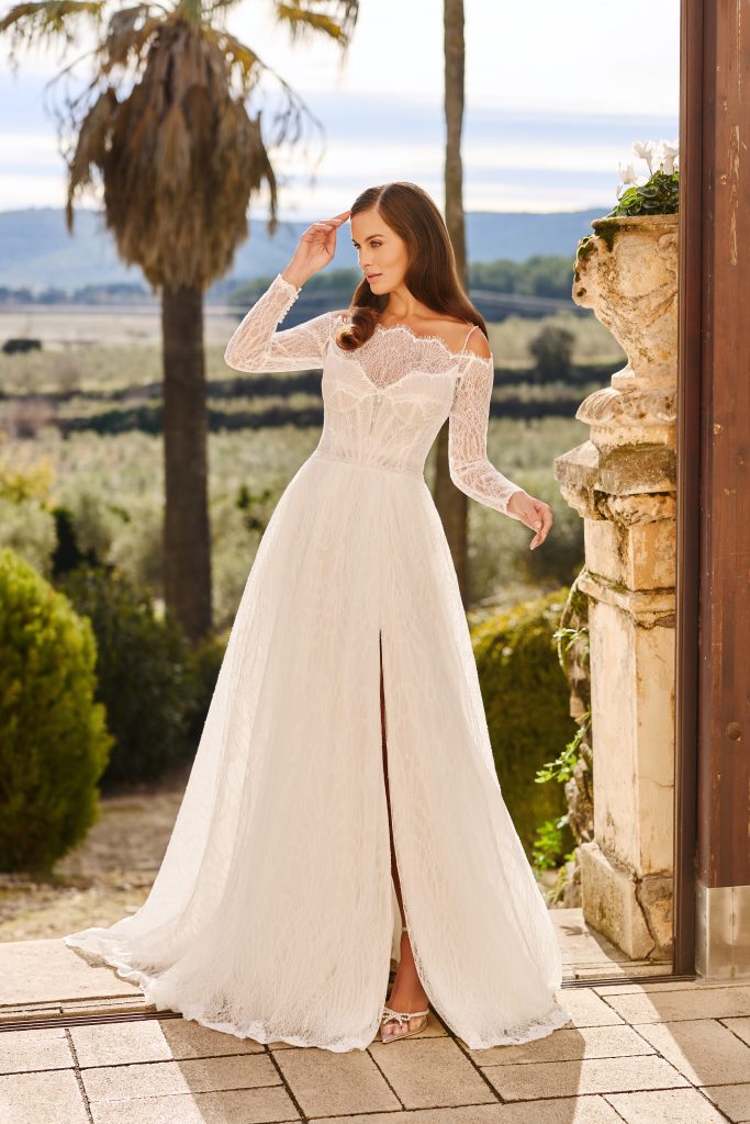 In questa foto, una modella posa all’aperto con un abito da sposa romantico in pizzo, con spalle scoperte e maniche lunghe. L’abito presenta uno spacco frontale che rivela le scarpe. Lo scatto fa parte della selezione di abiti da sposa invernali 2026 di Monica Loretti