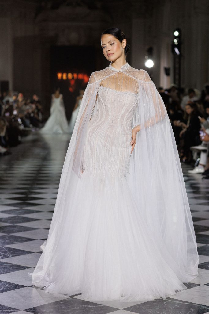 In questa foto, una modella sfila in passerella indossando un abito a sirena ricamato con perline e dettagli luminosi, completato da un mantello trasparente che cade leggero dalle spalle. L’immagine rappresenta perfettamente l’eleganza senza tempo degli abiti da sposa invernali 2026.