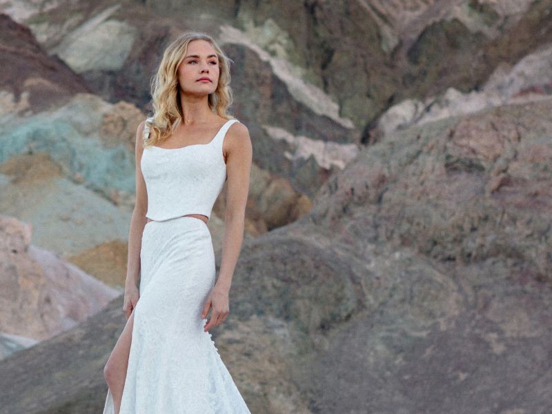 In questa foto, una modella posa all'aperto davanti a una scenografia rocciosa naturale, indossando un abito da sposa due pezzi composto da un top con spalline sottili e una gonna lunga con spacco laterale. Il tessuto ha una texture delicata e raffinata. Il look, essenziale e contemporaneo, rappresenta perfettamente lo stile degli abiti da sposa moderni 2026, pensati per spose che desiderano eleganza minimal e libertà di movimento.