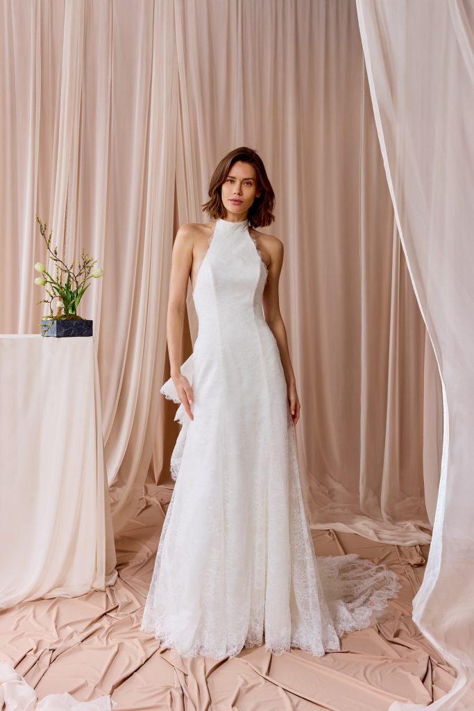 In questa foto, una modella posa davanti a un fondale rosa cipria indossando un abito da sposa moderno 2026 dallo stile minimal chic. Il vestito ha collo alto e taglio dritto, interamente in pizzo leggero, con dettagli raffinati lungo i bordi.