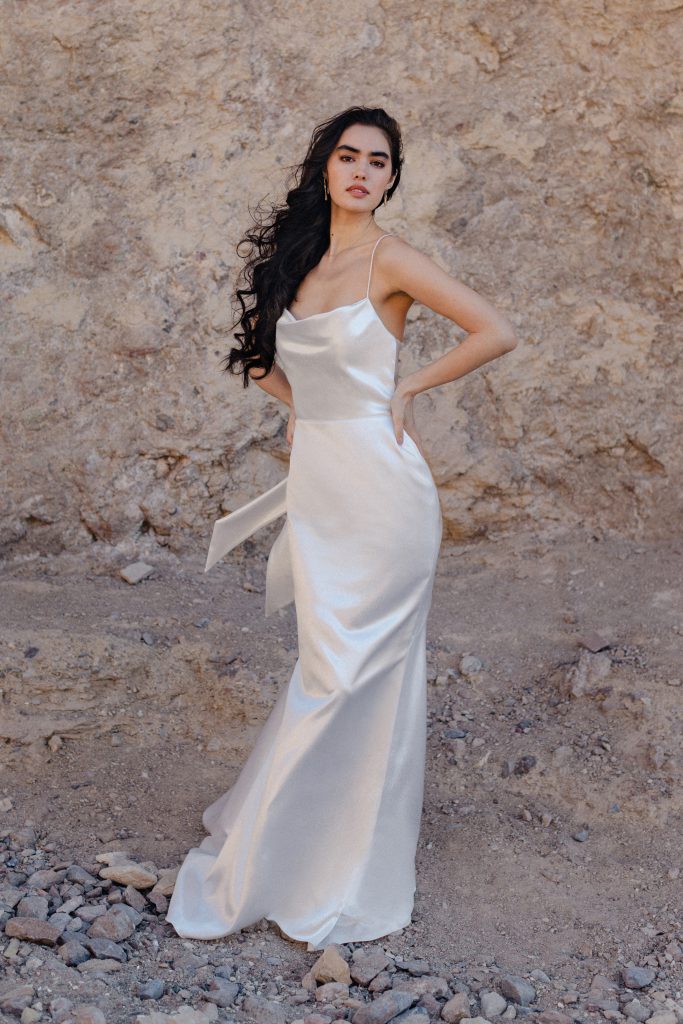 In questa foto, una modella posa davanti a una parete rocciosa indossando uno degli abiti da sposa moderni 2026: un vestito in raso lucido bianco perla con spalline sottilissime e scollo morbido. Il design è semplice, elegante e sensuale, perfetto per spose minimaliste e raffinate.