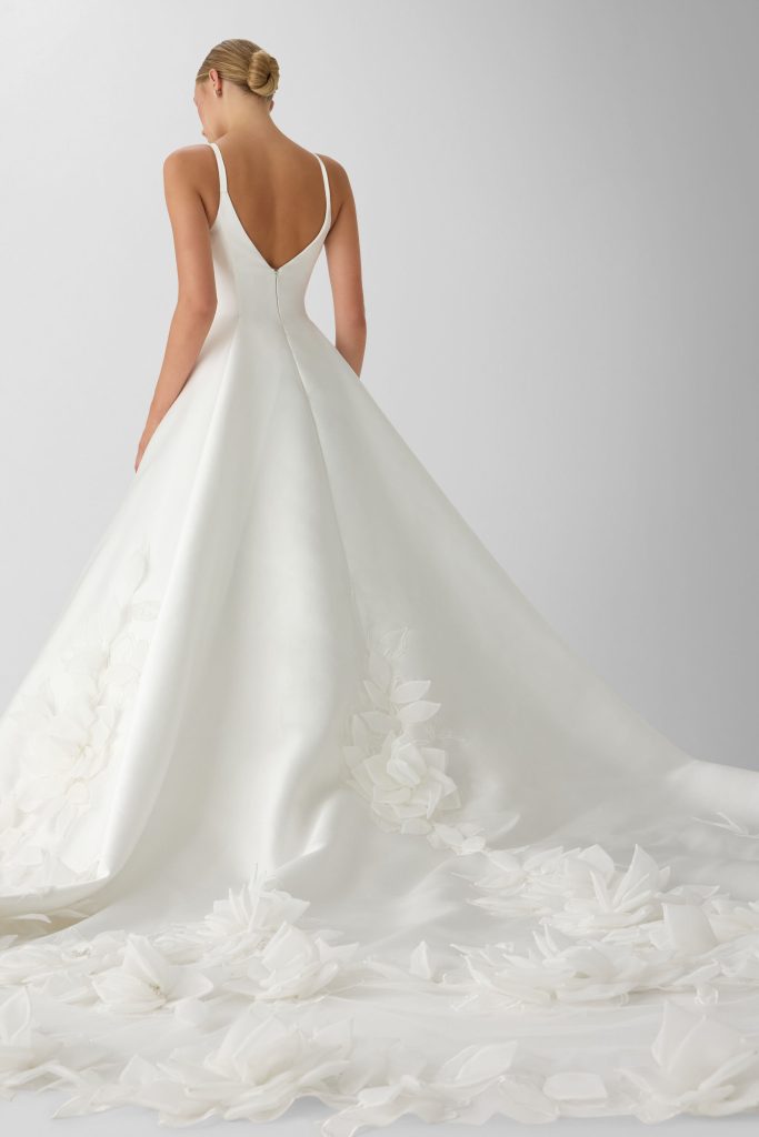 In questa foto, una sposa è ritratta di spalle mentre indossa un elegante abito da sposa moderno 2026 con scollo a V profondo e spalline sottili. Il vestito ha una lunga coda impreziosita da applicazioni floreali in rilievo tono su tono che danno movimento e raffinatezza al tessuto liscio.