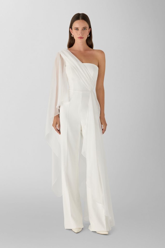 In questa foto, una modella con i capelli lisci porta uno dei più eleganti abiti da sposa moderni 2026: una tuta bianca con taglio monospalla. Il pantalone è ampio, mentre il bustino presenta un drappeggio fluido e un inserto in chiffon trasparente che cade con leggerezza da una spalla.