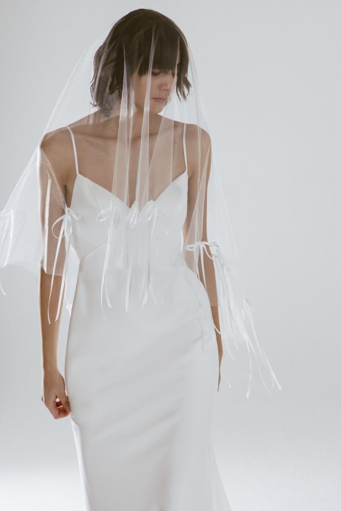 In questa foto, una sposa indossa uno degli abiti da sposa moderni 2026 più eterei e delicati. Il vestito, bianco latte e senza maniche, è completato da un velo lungo con fiocchi in tulle cuciti sul bordo. Il look è intimo, romantico e contemporaneo.