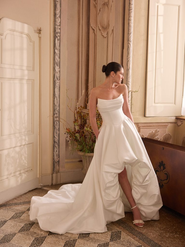In questa foto, la modella posa in un interno elegante indossando un abito da sposa moderno 2026 con corpetto strutturato e gonna ampia asimmetrica. Il modello è senza spalline e mette in mostra le gambe grazie a un’apertura centrale sulla parte frontale.