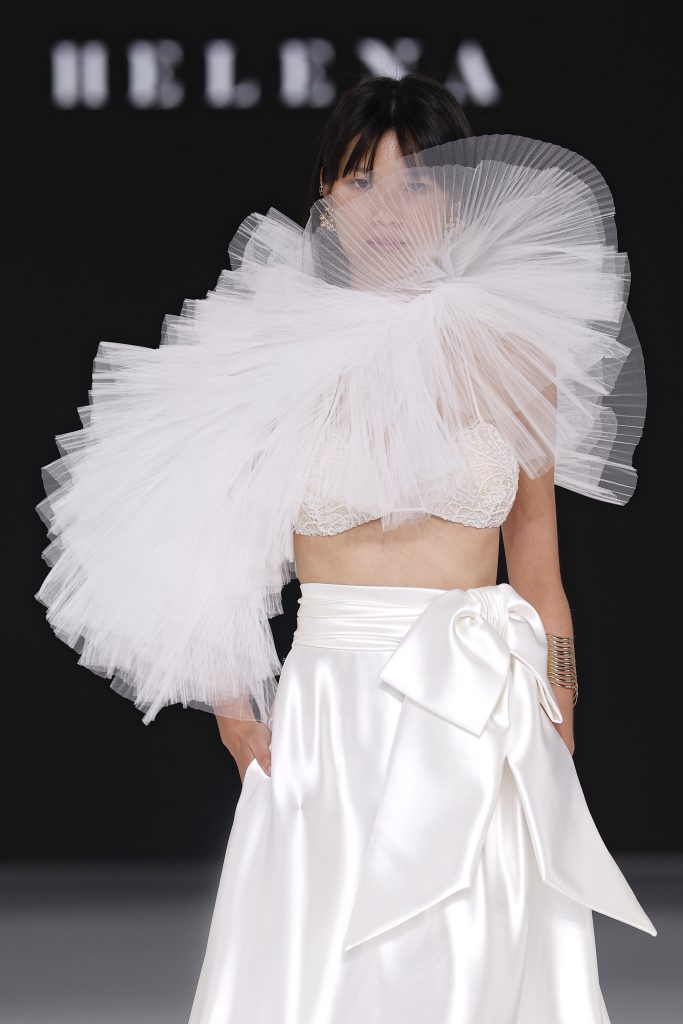 In questa foto, una modella sfila con un completo audace e scultoreo, tra gli abiti da sposa moderni 2026 più iconici. Il top è un reggiseno in pizzo, mentre sulla spalla destra campeggia un’enorme struttura in tulle a ventaglio. La gonna in raso ha un maxi fiocco laterale. Il risultato è un look d’alta moda, d’impatto e non convenzionale.
