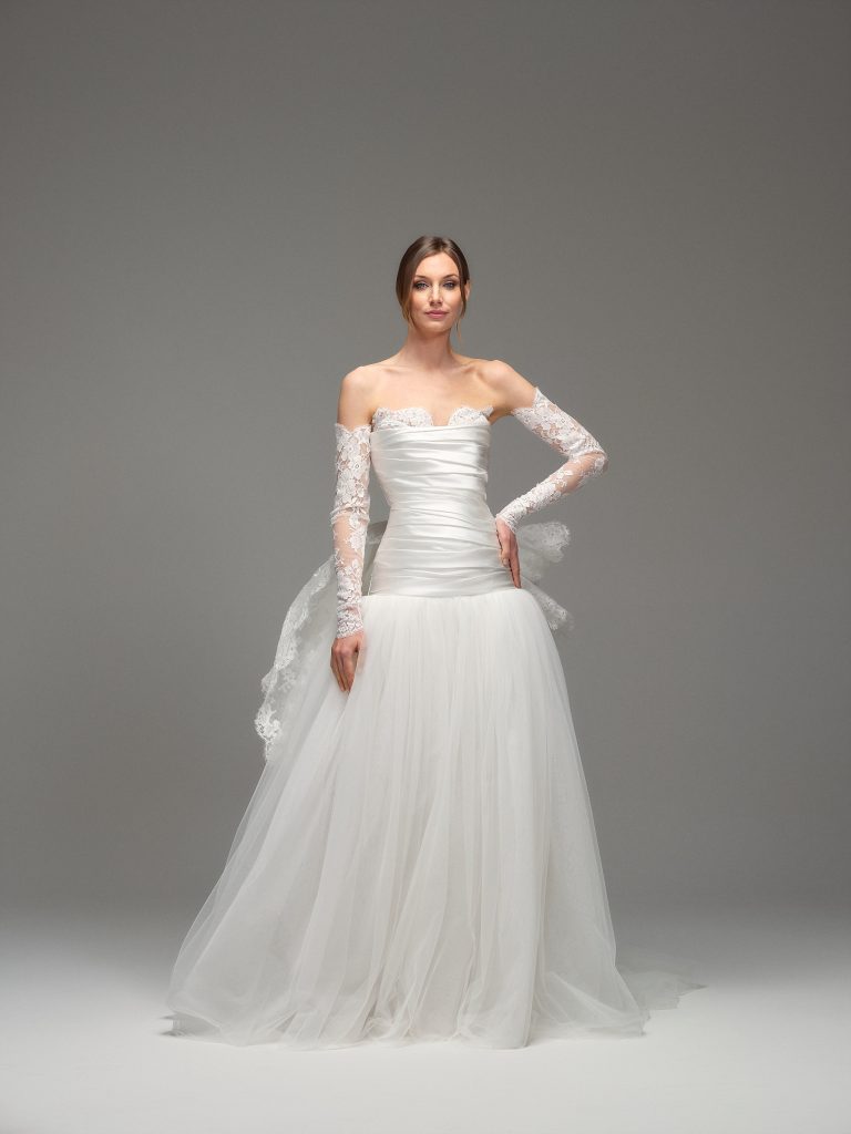 In questa foto, una modella indossa un abito da sposa moderno 2026 con corpetto drappeggiato, spalle scoperte e maniche in pizzo trasparente. La gonna è ampia e leggera, in tulle bianco, con un effetto vaporoso e romantico.