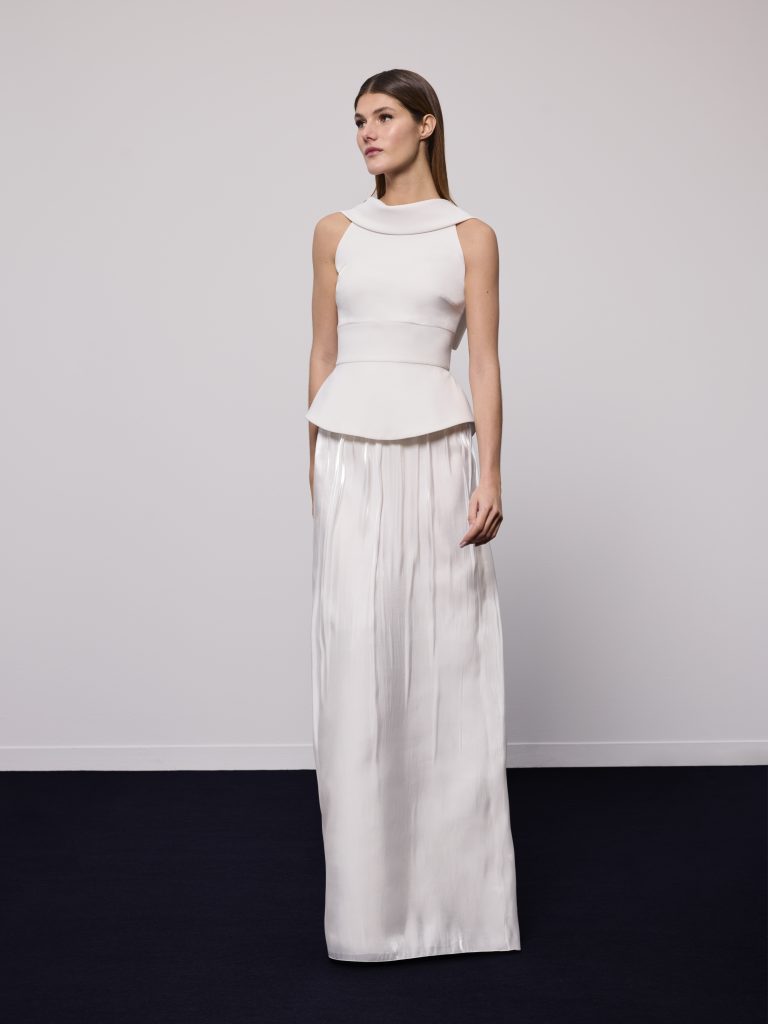 In questa foto, una modella sfoggia un abito da sposa moderno 2026 dal design essenziale e architettonico. Il top è strutturato con taglio peplo e girocollo ampio, mentre la gonna lunga e liscia cade morbida fino a terra con delicati riflessi.