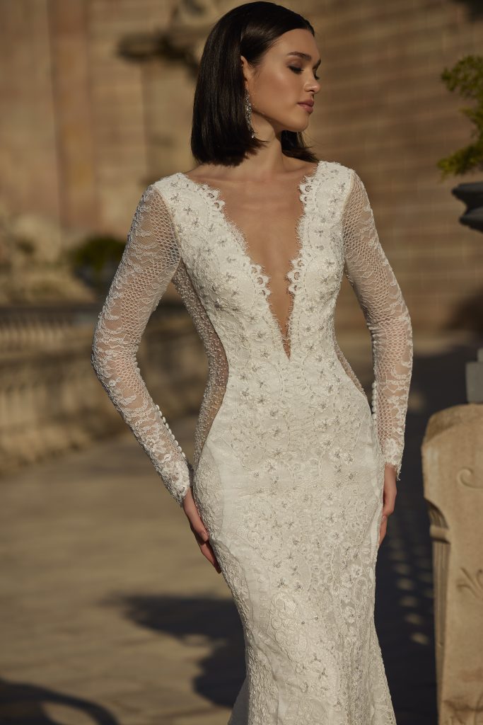 In questa foto, la modella indossa un abito da sposa moderno 2026 dal taglio a sirena con profondo scollo a V. Il vestito è in pizzo ricamato con maniche lunghe trasparenti e dettagli effetto rete che valorizzano la silhouette.