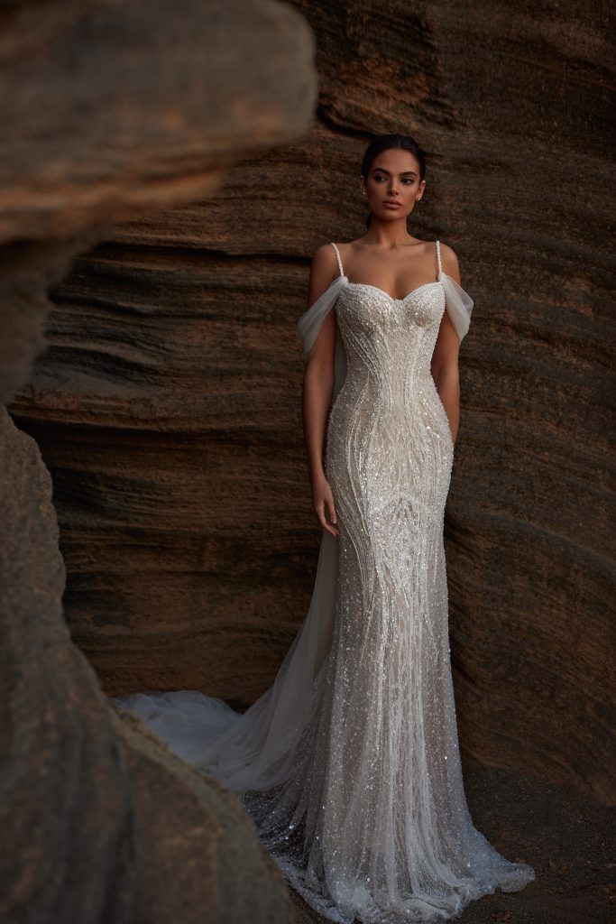 In questa foto, una modella indossa uno degli abiti da sposa particolari 2026 firmati Milla Nova: un vestito aderente con corpetto strutturato, decorazioni scintillanti e spalline sottili con tulle drappeggiato che scende sulle braccia. Lo sfondo naturale di rocce sabbiose crea un contrasto suggestivo con la lucentezza del tessuto.