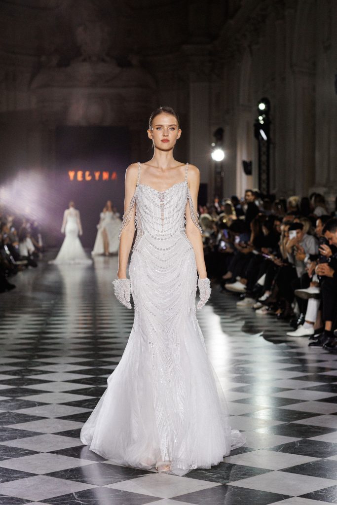 In questa foto, la modella sfila su una passerella con un abito da sposa particolare 2026 firmato Yedyna. Il vestito è ricco di perline e paillettes disposte in motivi ondulati, con sottili spalline e maniche gioiello pendenti. L’atmosfera elegante della sfilata esalta la raffinatezza dell’abito.