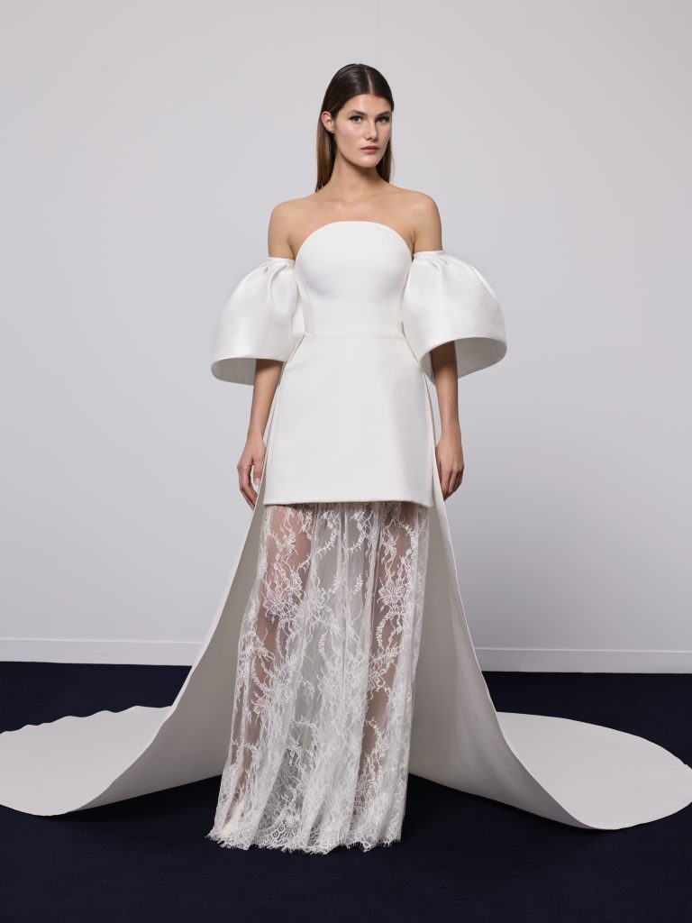 In questa foto, una modella indossa un abito da sposa particolare 2026 dal design innovativo: la parte superiore ricorda una minigonna con maxi maniche balloon, mentre la parte inferiore è composta da una gonna lunga trasparente in pizzo. Il look è pulito ma d’impatto, perfetto per chi cerca uno stile non convenzionale.