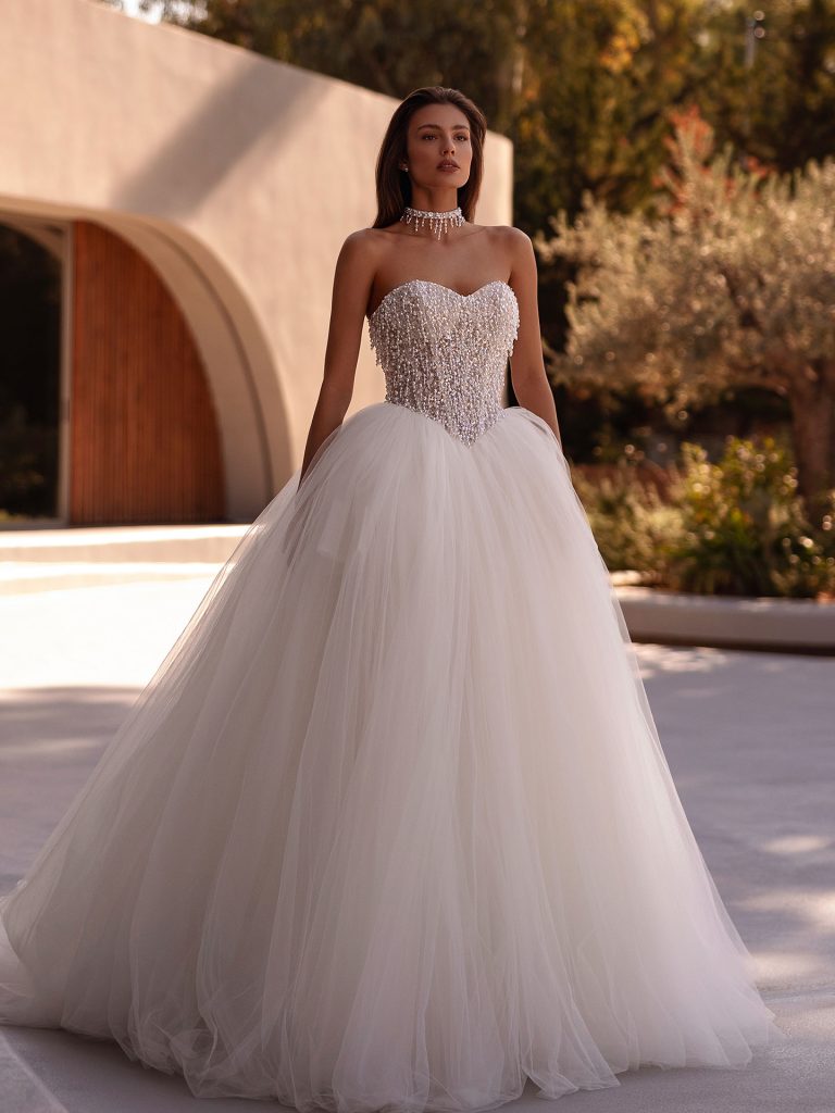 In questa foto, una sposa indossa un abito da ballo con corpetto interamente ricamato di cristalli e perline, abbinato a una voluminosa gonna in tulle. L’immagine, scattata all’aperto in una location moderna, celebra l’eleganza brillante degli abiti da sposa particolari 2026.