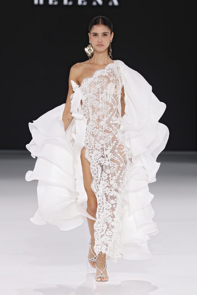 In questa foto, una modella sfila indossando un abito da sposa particolare 2026 con un design audace: un solo spallaccio, ampissimi volant bianchi asimmetrici e un corpo in pizzo trasparente ricamato con motivi floreali. Lo spacco profondo e le trasparenze esaltano il look scenografico e teatrale.