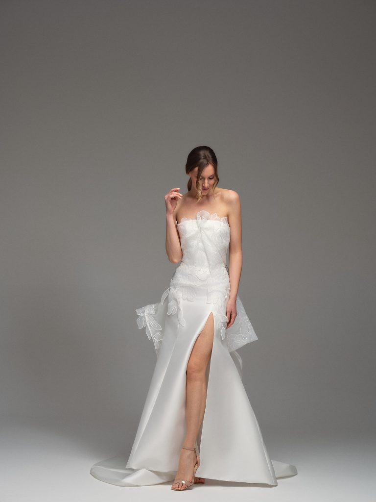 In questa foto, una sposa posa con eleganza in uno studio neutro, indossando un abito da sposa particolare 2026 con corpino senza spalline e applicazioni floreali tono su tono. La gonna presenta un profondo spacco frontale, dettagli in rilievo e una struttura scultorea che unisce romanticismo e modernità.