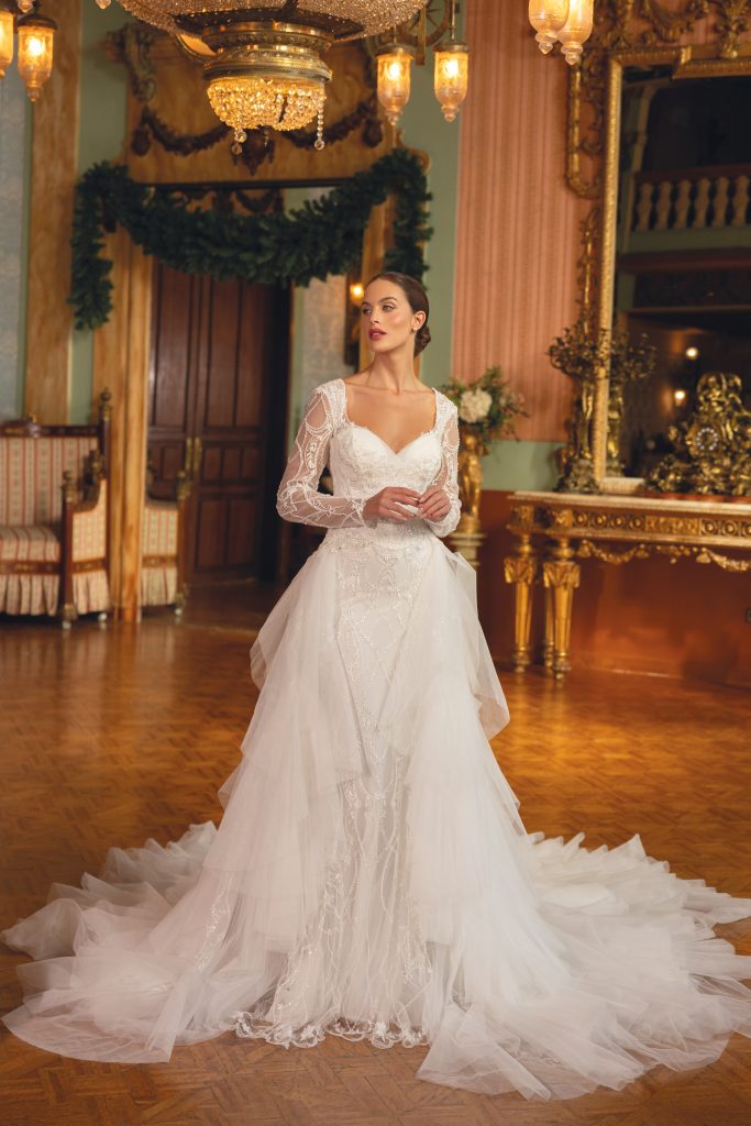 In questa foto, una sposa posa in un sontuoso salone illuminato da lampadari antichi. L’abito, in stile romantico, ha maniche lunghe in pizzo, dettagli gioiello e una gonna ampia con strati di tulle. Una scelta perfetta tra gli abiti da sposa particolari 2026 per chi sogna un matrimonio fiabesco.