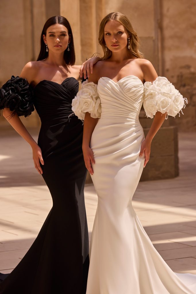 In questa foto, due modelle posano affiancate indossando abiti da sposa particolari 2026 con maniche voluminose a forma di fiori: una in total black, l’altra in bianco. Entrambi gli abiti sono aderenti, con scollo a cuore e taglio a sirena, perfetti per spose glamour che vogliono osare.