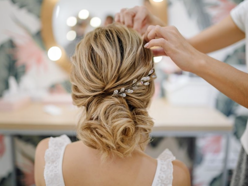 in questa foto si vede un'acconciatura sposa capelli lunghi raccolti di colore biondo chiaro. la sposa è seduta mentre un parrucchiere sposa ultima l'acconciatura