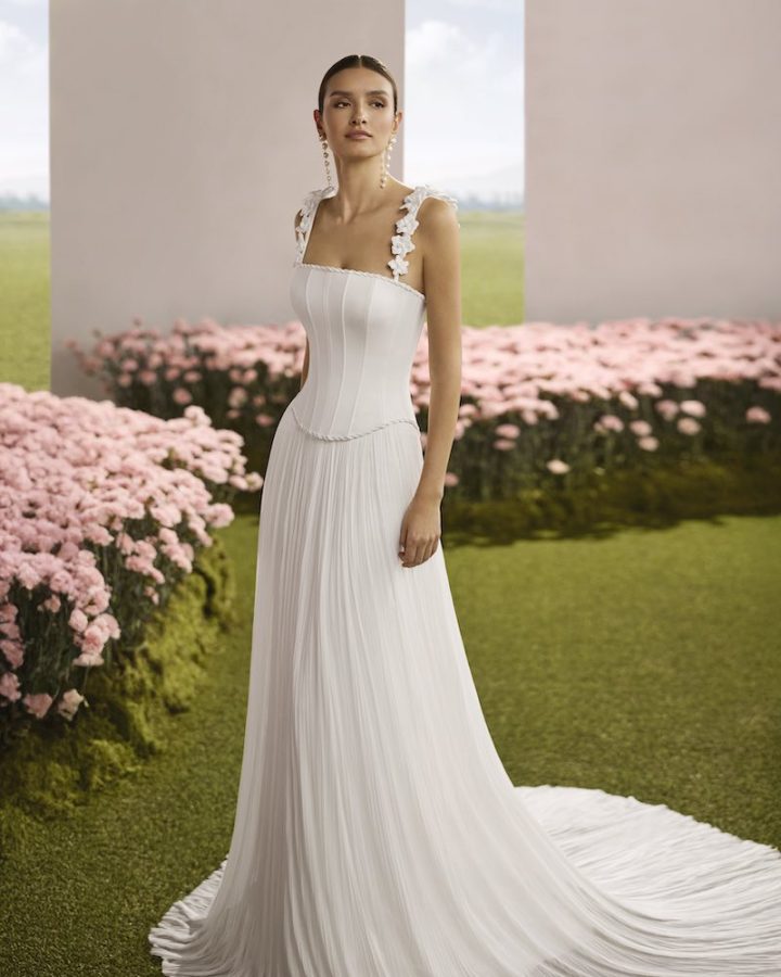 In questa foto, una modella posa indossando un abito della collezione sposa Rosa Clará Couture 2026 con taglio scivolato, corpetto a vita basca e bretelle con applicazioni floreali 3D