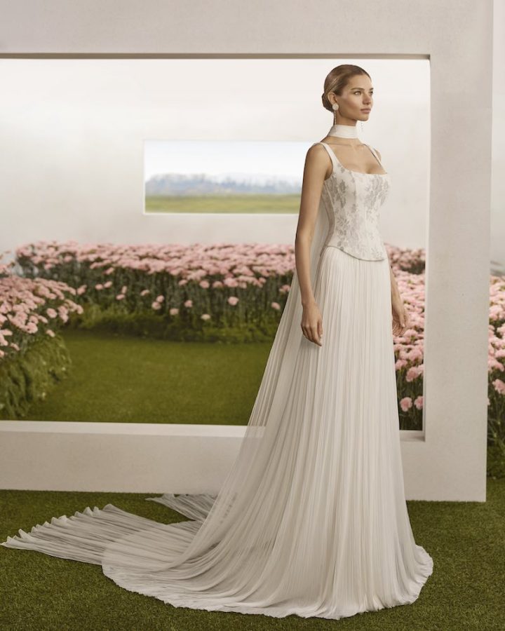 In questa foto, una modella posa indossando un abito della collezione sposa Rosa Clará Couture 2026 con taglio scivolato, corpetto a vita basca ricamato con fili d'argento