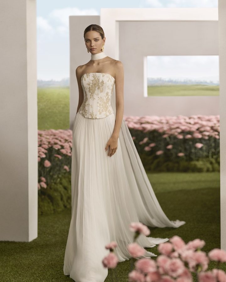 In questa foto, una modella posa indossando un abito della collezione sposa Rosa Clará Couture 2026 con taglio scivolato, corpetto a vita basca ricamato con fili dorati