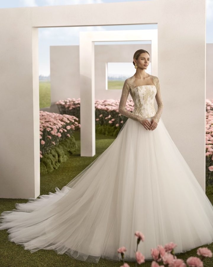 In questa foto, una modella posa indossando un abito della collezione sposa Rosa Clará Couture 2026 con taglio principessa, corpetto a vita basca ricamato con fili oro, coprispalla abbinato e gonna in tulle