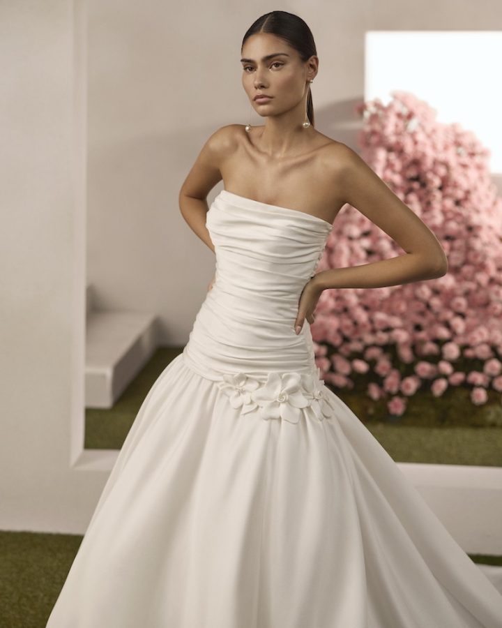 In questa foto, una modella inquadrata a tre quarti posa indossando un abito da sposa con corpetto drappeggiato e fiori 3D in in vita